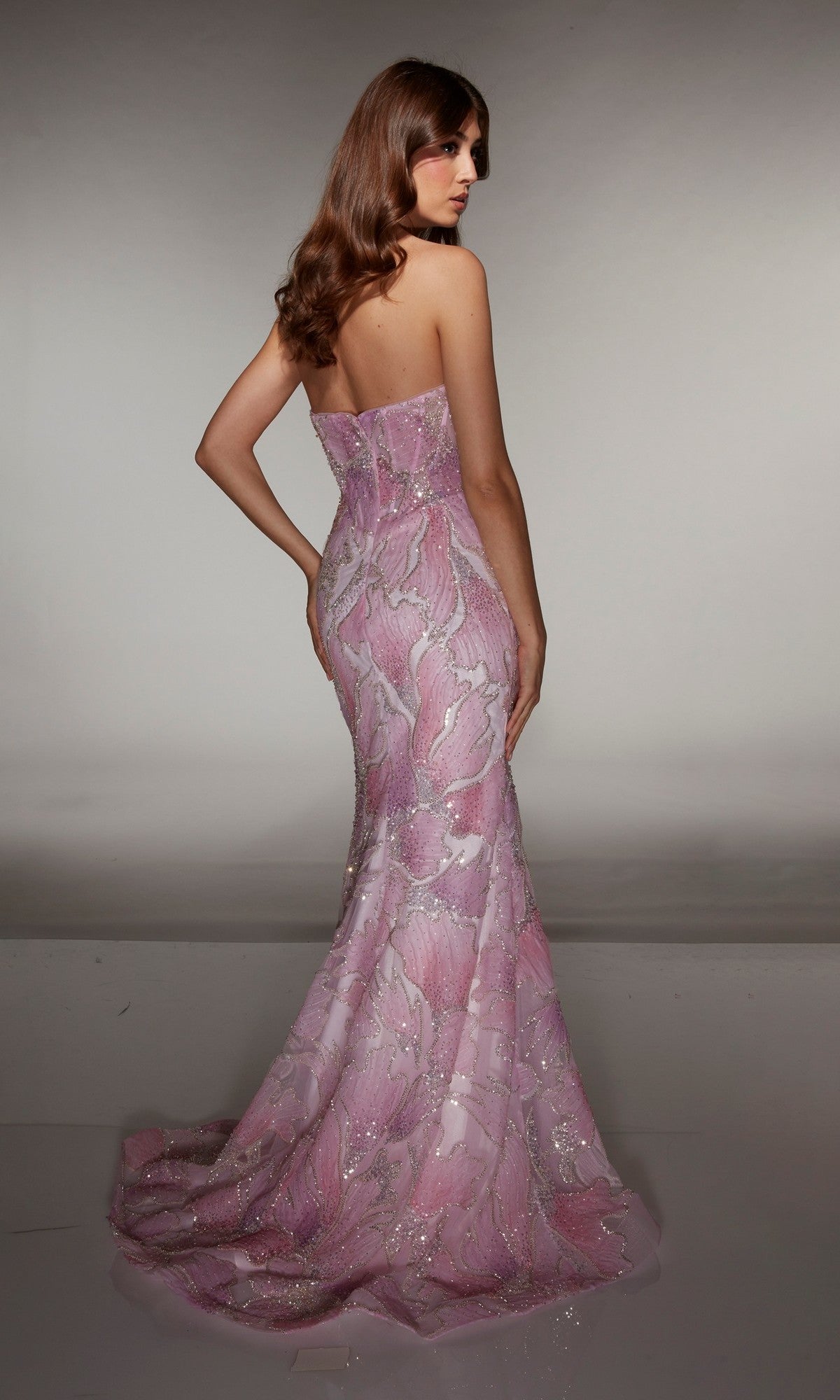 Person standing in a studio wearing the Alyce 62049 long prom dress with a strapless floral design, sheer corset bodice, floor-length skirt, and sparkling details, elegantly looking over their shoulder.